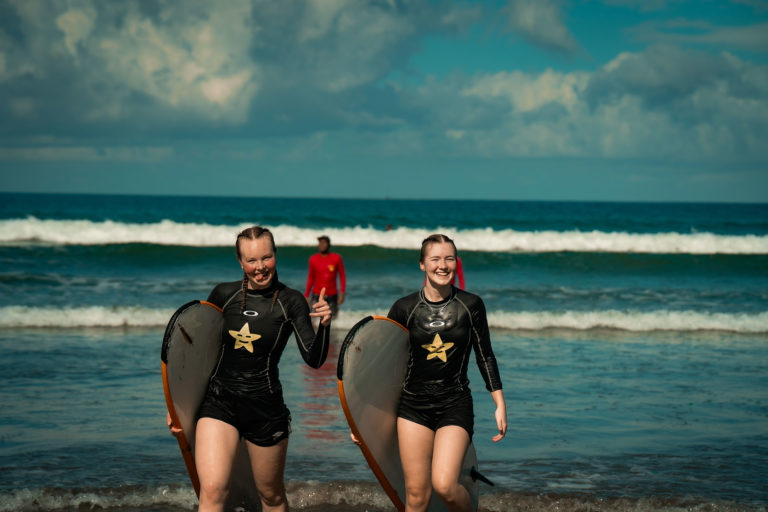 To jenter med surfebrett, ene smiler, andre skjærer grimase og viser tommel opp